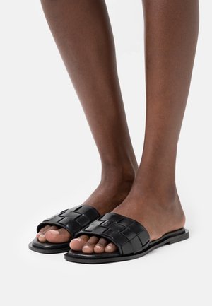 Sandales à lanières en cuir noir avec un design tissé, semelle plate et style ouvert aux orteils, mises en avant sur une paire de jambes.