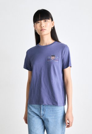 T-shirt en coton violet avec col rond, manches courtes et logo GANT brodé sur la poitrine, associé à un jean bleu clair.