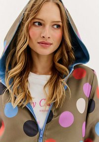 Kurtka z kapturem w brązowej bazie, zdobiona dużymi, wielokolorowymi kropkami. Posiada niebieską podszewkę i zamek z przodu.