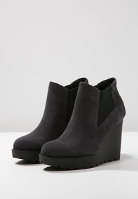 Stivaletti alla caviglia in suede nero con zeppa, pannelli laterali in elastico, punta rotonda e suola texture, caratterizzati da una finitura liscia e un design moderno.