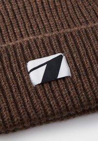 Bonnet en tricot côtelé marron avec une étiquette noir et blanc présentant un design géométrique. Le matériau semble doux et texturé.