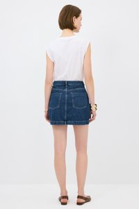 Dżinsowa spódnica mini z wysokim stanem, dwoma tylnymi kieszeniami i surowym wykończeniem. Noszona z białą topem bez rękawów i płaskimi brązowymi sandałami.