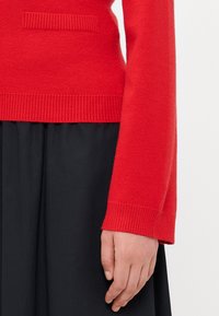Pull en tricot rouge avec ourlet et manches côtelés, doté d'une petite poche texturée à l'avant. Associé à une jupe noire.