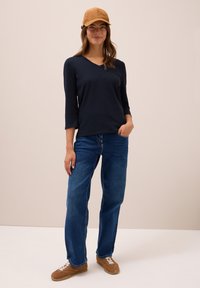 Marineblaues Oberteil mit drei Viertelärmel, blaue gerade Jeans und beige Wildleder-Baseballmütze. Braune Sneaker mit weißen Schnürsenkeln runden das Outfit ab.