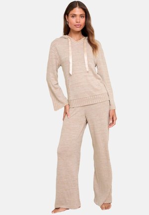 Gaiši bež loungewear komplekts, kas sastāv no kapuces augšdaļas ar garām piedurknēm un satīna auklām, kas apvienots ar platām biksēm no līdzīgas tekstūras.