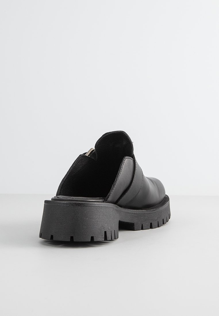 Chaussure noire en cuir glissante avec un petit talon épais et une semelle texturée et striée. Présente un dessus lisse avec des détails de couture minimaux.