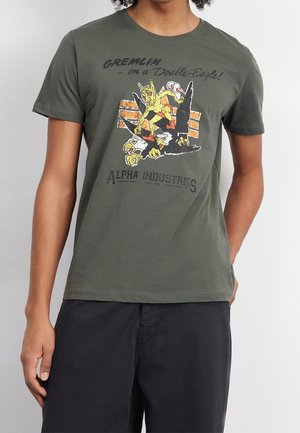 Olivgrünes Baumwoll-T-Shirt mit einem grafischen Aufdruck eines Gremlins auf einem Flugzeug, mit gelben und roten Akzenten. Kurze Ärmel und Rundhalsausschnitt.