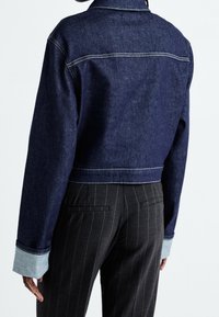 Kortsiktig denimjacka i mörkblått med mjuk textur, som har ljusblåa manschetter och kontrasterande vit sömnad. I kombination med randiga byxor.