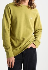 Olivově zelená mikina vyrobená z měkké tkaniny, s kulatým výstřihem, dlouhými rukávy a malým logem "Obey" na hrudi. Viditelné černé kalhoty.