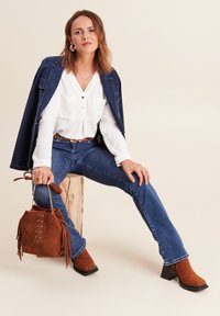 Bottines en suède marron, jeans bleus, blouse blanche à boutons, veste en denim, sac à main marron avec franges et clous, posés sur un bloc en bois.