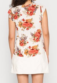Top floral en blanco con patrones en rojo, naranja y púrpura. Presenta mangas cortas con volantes y un dobladillo redondeado, combinado con una falda blanca.