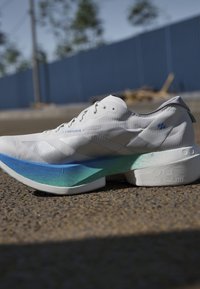 Witte hardloopschoen met blauw-groene verloopzool die op asfalt rust met een blauwe omheining en bomen op de achtergrond.
