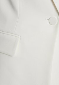 Blazer bianco con una texture liscia, vestibilità su misura e un bottone circolare singolo come accentuazione sulla tasca anteriore.