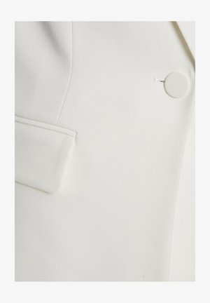 Blazer blanc avec une texture lisse, une coupe élégante et un accent de bouton circulaire unique sur la poche avant.