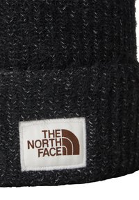 Berretto nero lavorato a maglia con un motivo testurizzato. Presenta un'etichetta bianca con "THE NORTH FACE" in marrone e un logo ad arco. Materiale morbido e caldo.
