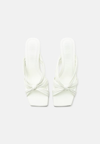 Ζεύγος λευκών σανδαλιών με ανοιχτά δάχτυλα και τετράγωνες μύτες, λεπτά πολλαπλά λουράκια δεμένα μπροστά και στυλ slip-on σε λευκό φόντο.