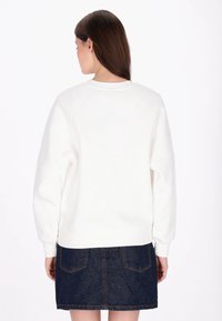 Fehér pulóver laza szabással, bordázott mandzsettákkal és szegéllyel. Sötét farmer szoknyával párosítva, melyen hátsó zsebek és varrási részletek találhatók.