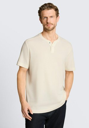 Mann mit kurzen lockigen Haaren und Bart, der ein cremefarbenes Henley-Shirt mit kurzen Ärmeln und dunkle Hose trägt, steht mit einer Hand in der Tasche.
