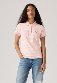 Polo a righe in rosa chiaro con colletto, maniche corte e una piccola etichetta con logo sul petto. Abbinato a jeans strappati.