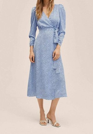 Femme portant une robe portefeuille à manches longues bleu clair avec des pois noirs et des sandales à talons blanches à bouts ouverts, sur un fond neutre.