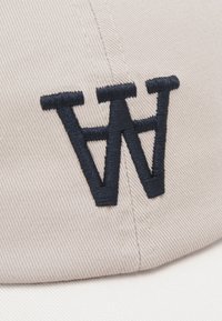 Căciulă de baseball din bumbac bej, cu un logo brodat "W" de culoare albastră închisă pe partea din față. Prezintă un material texturat și un design structurat.