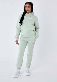 Hoodie verde chiaro con tasca frontale e logo bianco, abbinato a pantaloni della tuta coordinati. Il materiale sembra morbido con una vestibilità comoda e caviglie elastiche.