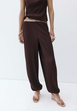 Mujer vestida con pantalones anchos de pierna con textura acanalada en marrón oscuro, top sin mangas a juego, sandalias con cuña transparentes y una cadena de cuentas en la cintura sobre fondo blanco.