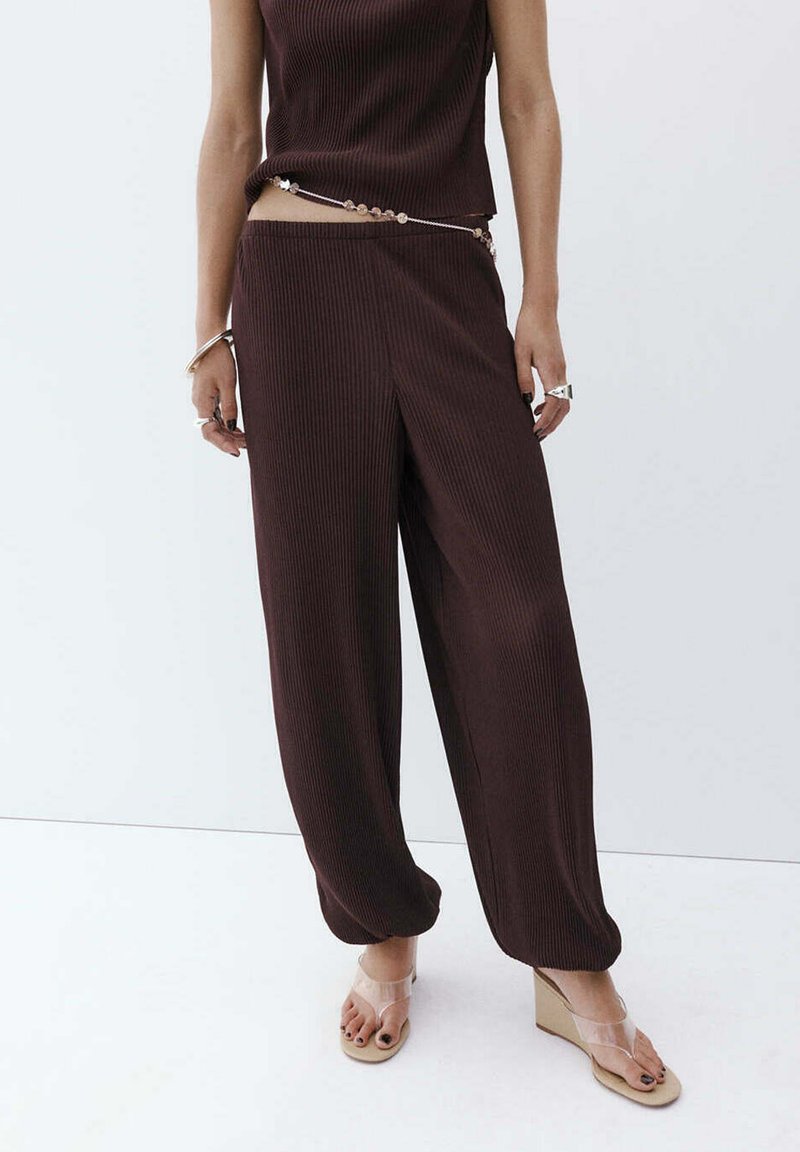 Femme portant un pantalon large côtelé marron foncé, un top sans manches assorti, des sandales compensées transparentes et une chaîne de taille perlée sur fond blanc.