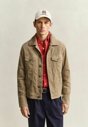 Young man wearing a beige jacket over a red shirt, navy pants, and a white baseball cap with embroidered text.