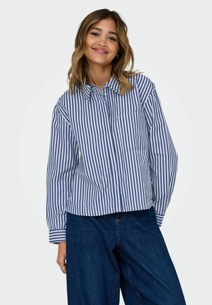 Camicia a righe con strisce verticali blu e bianche, colletto, taschino frontale e maniche larghe. Abbinata a pantaloni in denim.