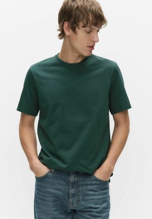 Junger Mann mit hellbraunen Haaren, der ein grünes, kurzärmeliges T-Shirt und blaue Jeans trägt, die Hände in den Taschen, schaut nach links.