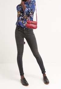 Blommig blå blus, svarta skinny jeans, uppvikta ärmar och svarta skor. Röd crossbody-väska ger kontrast. Slätt tyg, figurnära design.