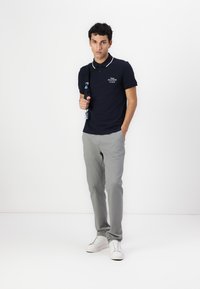 Marineblå polo med hvit trim, logo detaljer på brystet, kombinert med lysegrå bukser og hvite joggesko, stående foran en ensfarget bakgrunn.