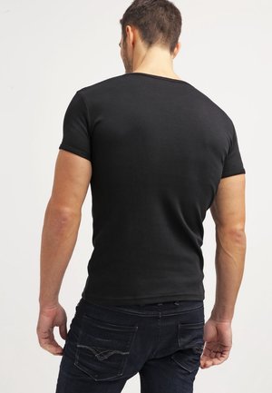 Mann trägt ein eng anliegendes schwarzes T-Shirt mit kurzen Ärmeln und dunkle Jeans, von hinten vor schlichtem hellem Hintergrund gesehen.