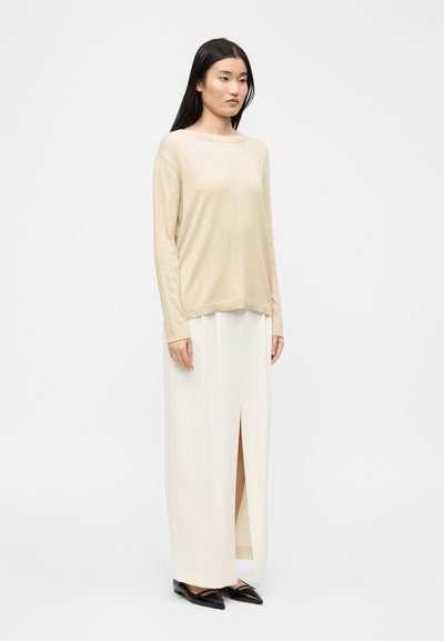 Femme aux longs cheveux noirs portant un pull beige côtelé à manches longues, une jupe longue blanche fendue haute, et des ballerines noires pointues, debout devant un fond blanc.