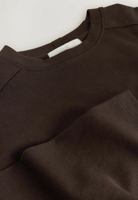 Dunkelbraunes Langarmshirt mit weicher, glatter Textur und Rundhalsausschnitt, ausgestattet mit einer kleinen Tasche auf der Vorderseite und minimalen Nahtdetails.