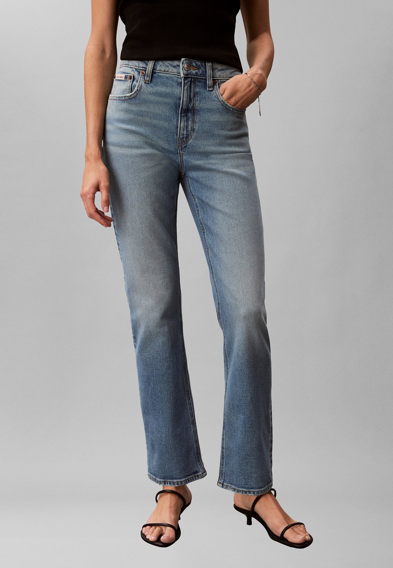 Femme portant un jean bleu délavé à jambes droites, un haut noir sans manches et des sandales à talons noires, debout avec une main dans la poche.