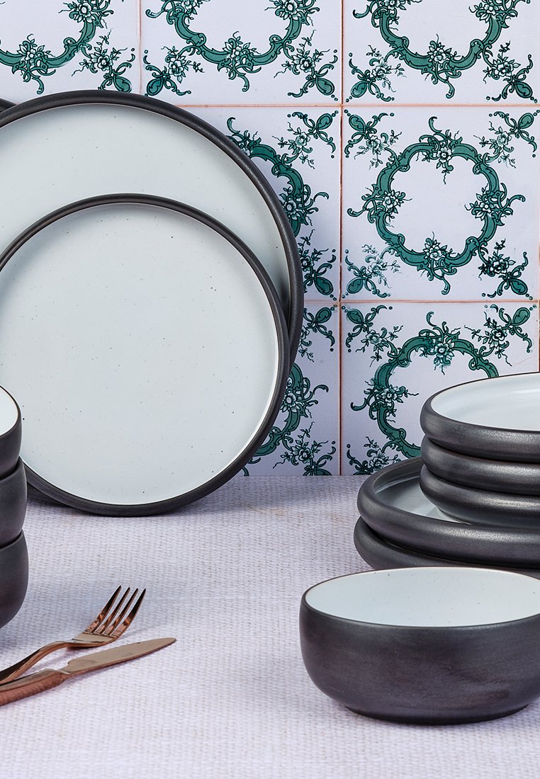 Ceramiczny zestaw naczyń z gładkimi białymi powierzchniami i ciemnoszarymi rantami. Zawiera talerze i miski ułożone na teksturowanej powierzchni.