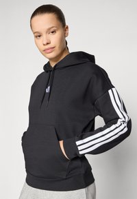 Sweatshirt preto com capuz e bolso frontal, apresentando detalhes em branco com três riscas nas mangas e um pequeno logo no peito.
