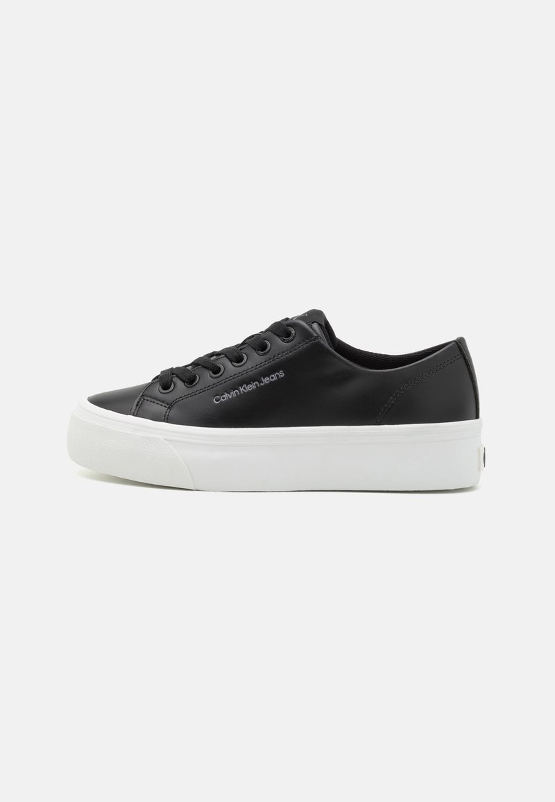 Sorte faux skinn sneakers med hvit gummisåle, rund tå, seks hull snøring, og "Calvin Klein Jeans" logo på siden.