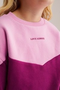 Rozā un violeta krāsu kombinācijas sporta krekls ar ribotu apkakli. Uz krūtīm ir izšūts uzraksts "MĪLESTĪBA VIENMĒR" atbilstošā krāsā.