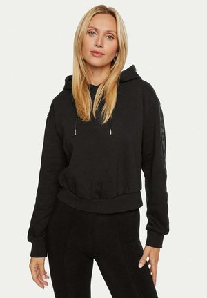 Felpa corta nera in tessuto morbido, con cappuccio regolabile tramite coulisse e dettagli con logo laterale. Abbinata a leggings neri coordinati.