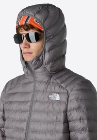 Grå isolerad huva-jacka med kviltad textur och kontrasterande dragkedja. Har logotyp på bröstet och en färgglad mönstrad mössa under.