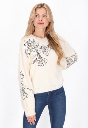 Cream sweater with floral embroidery in black and red, textured knit, relaxed fit, and wide sleeves, paired with blue jeans.