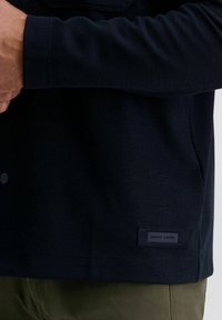 Navy-blaues, strukturiertes Langarmhemd mit einem geknöpften Kragen, das ein Etikett mit "pierre cardin" am Saum trägt. Über grü fabric getragen.