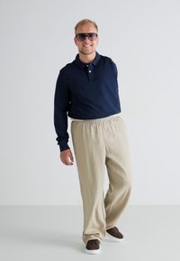 Polo shirt cu mânecă lungă de culoare bleumarin, pantaloni bej cu talie elastică și adidași maro. Persoana are părul scurt de culoare blondă și poartă ochelari de soare.