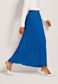 Frau, die in einem leuchtend blauen gefälteten Maxikleid, einer weißen Bluse und weißen Slingback-High-Heels vor einem schlichten hellen Hintergrund geht.