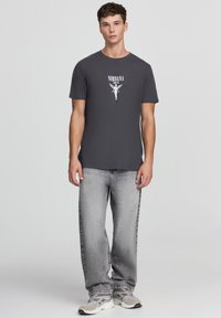 T-shirt in cotone grigio con grafica bianca "NIRVANA" e design di angelo. Indossato con jeans grigi chiari e sneaker grigie.