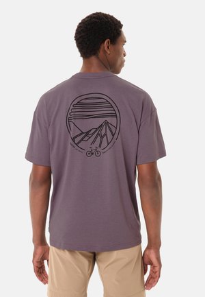 Uomo di spalle che indossa una maglietta viola con grafica nera di montagna e bicicletta e pantaloncini color kaki su sfondo bianco.