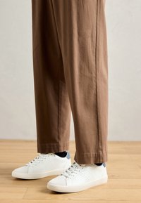 Personne portant un pantalon marron et des baskets blanches avec des chaussettes noires, se tenant sur un sol en bois contre un mur uni.